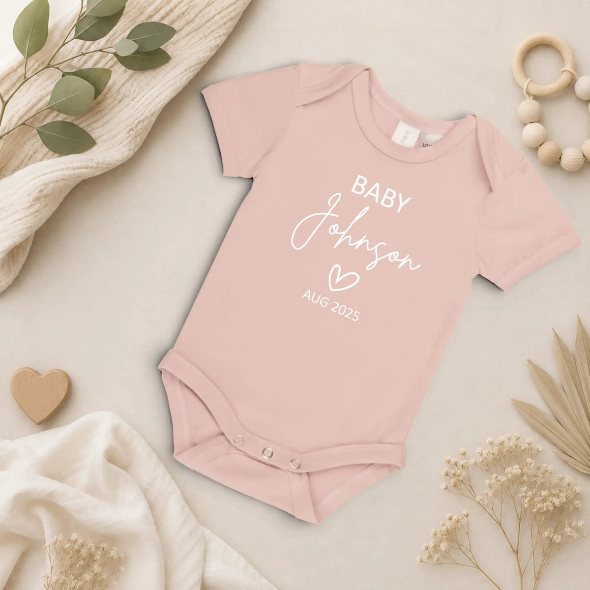 Blush Pink baby onesie with 'Baby Johnson' and 'AUG 2023' text, surrounded by decorative elements on a light background.
