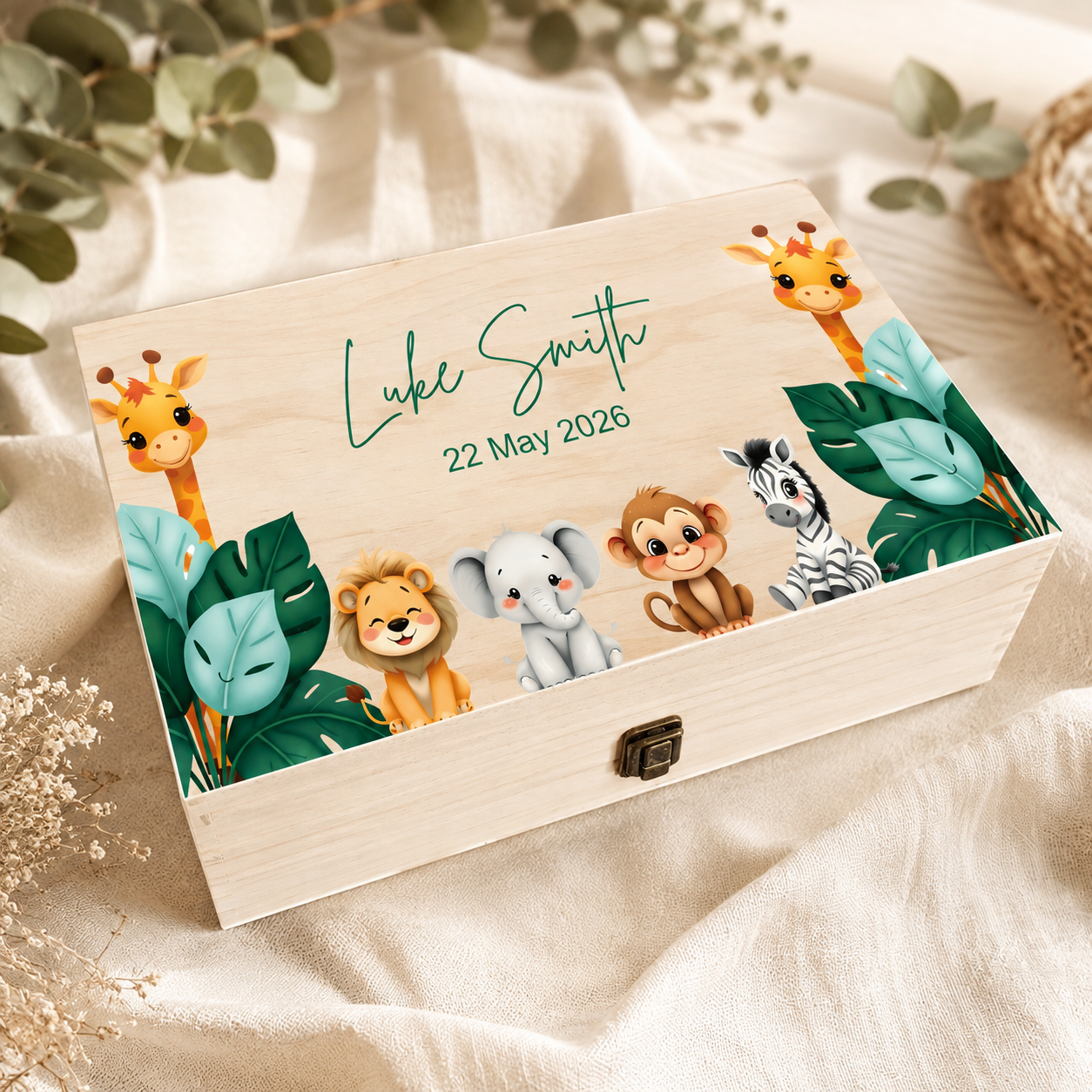 A personalized wooden keepsake box with a hinged lid, decorated with cartoon safari animals and the text 'Luke Smith 22 May 2026'. The box is styled on a beige fabric background with dried flowers and green leaves.