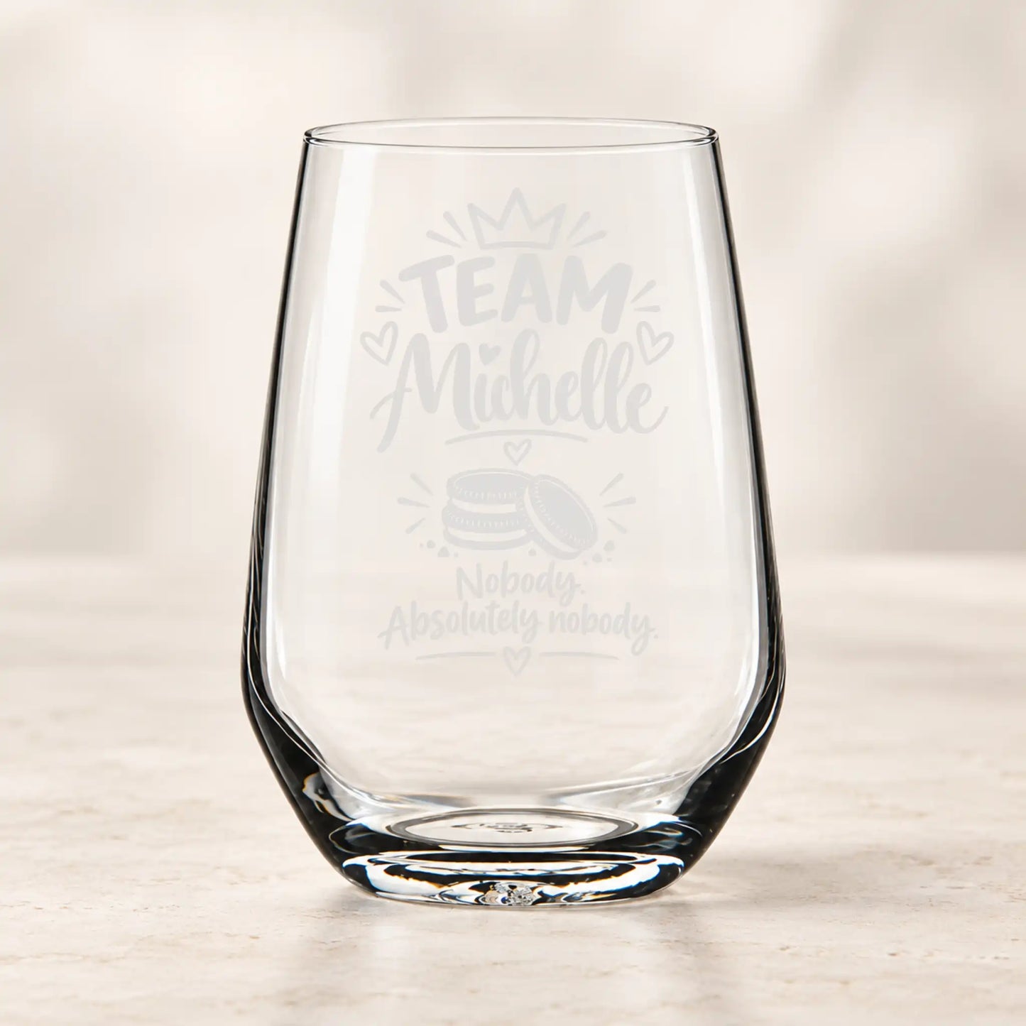 A clear stemless wine glass with custom light gray laser engravings that read 'TEAM Michelle' and 'Nobody Absolutely nobody' with a small cake graphic, set on a light marble surface against a soft beige background.