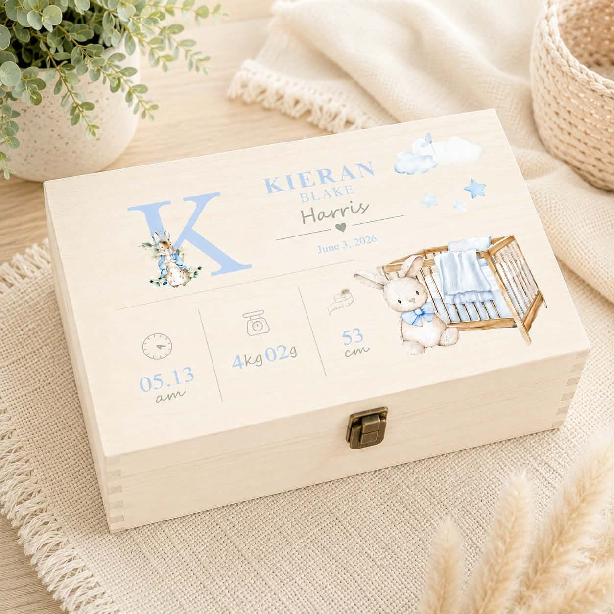 A personalized wooden baby keepsake box with a metal clasp, decorated with blue and white illustrations of a bunny, clouds, stars, and baby details, resting on a beige woven mat with a plant and blanket in the background.