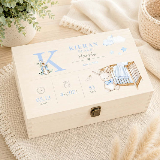 A personalized wooden baby keepsake box with a metal clasp, decorated with blue and white illustrations of a bunny, clouds, stars, and baby details, resting on a beige woven mat with a plant and blanket in the background.