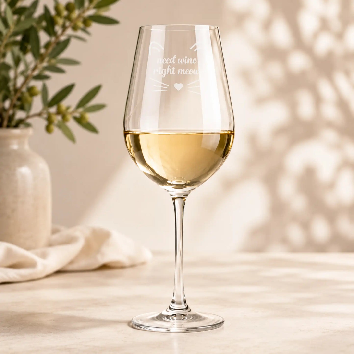 A clear wine glass with a stem, partially filled with white wine, standing on a light tabletop. Etched near the bowl is the phrase 'need wine right meow' with small whisker and heart details.
