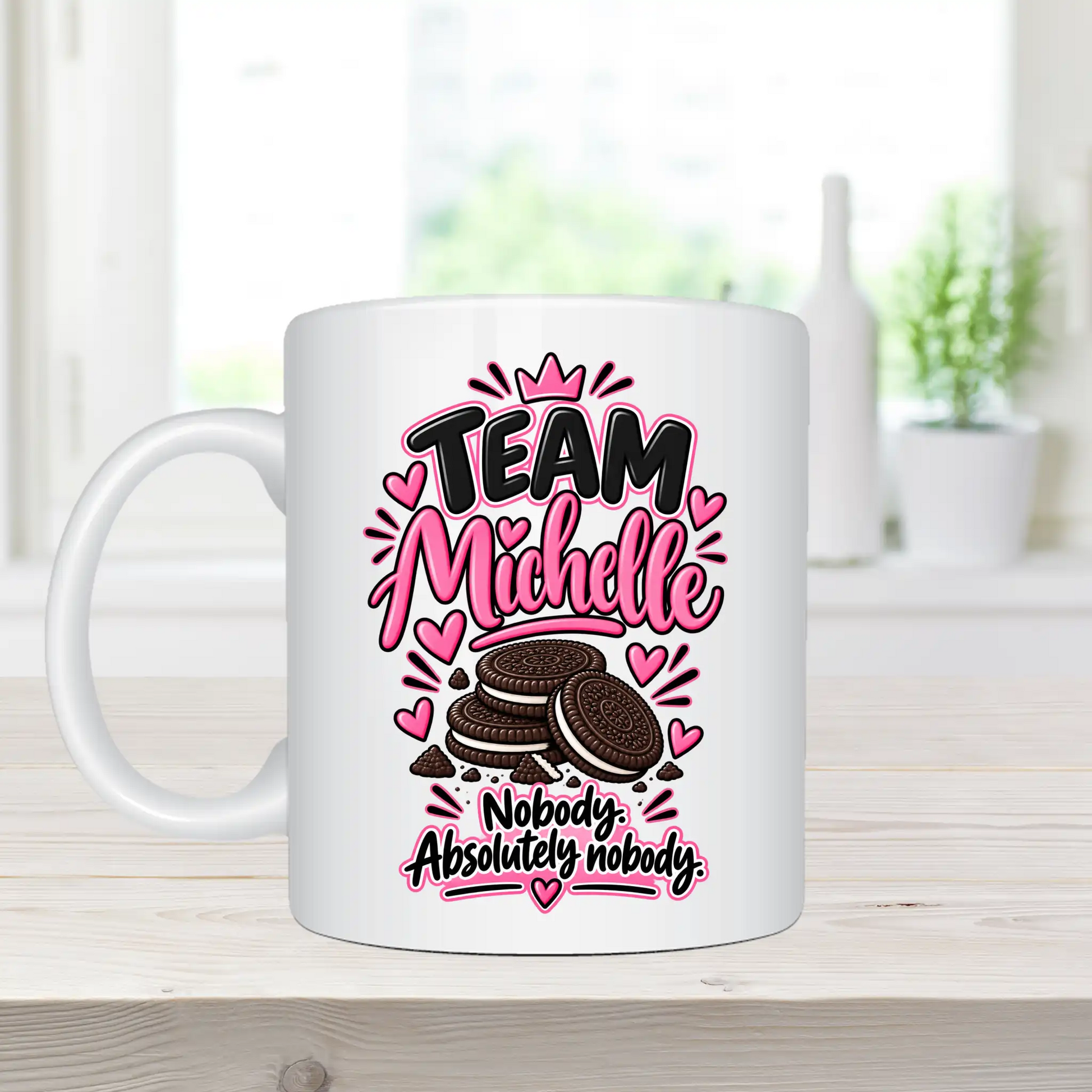 A white ceramic coffee mug with a custom graphic design, placed on a light wooden tabletop with a blurred kitchen background. The graphic features the text 'TEAM Michelle' in pink and black, decorated with pink hearts and a small crown, above an illustration of chocolate sandwich cookies.