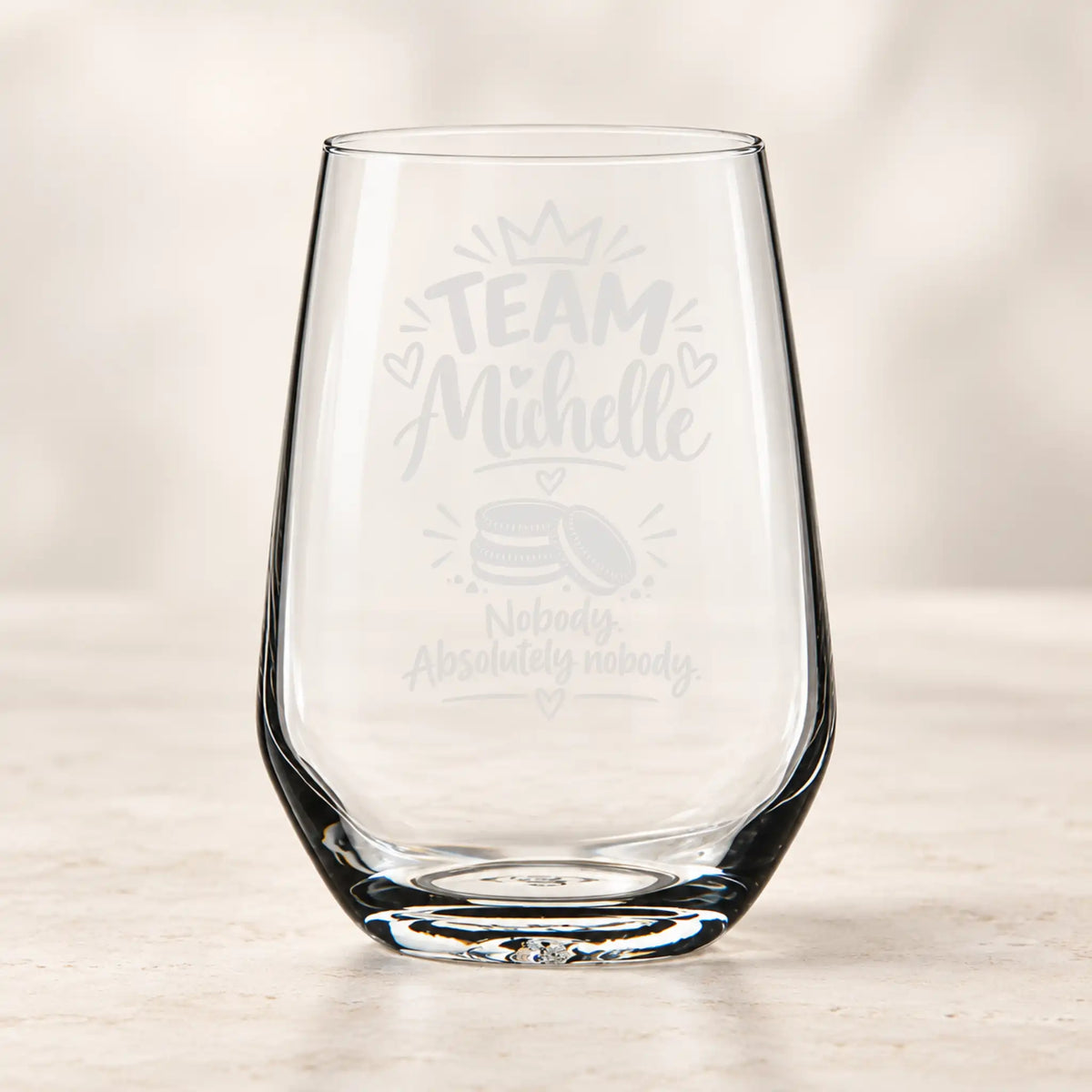 A clear stemless wine glass with custom light gray laser engravings that read 'TEAM Michelle' and 'Nobody Absolutely nobody' with a small cake graphic, set on a light marble surface against a soft beige background.