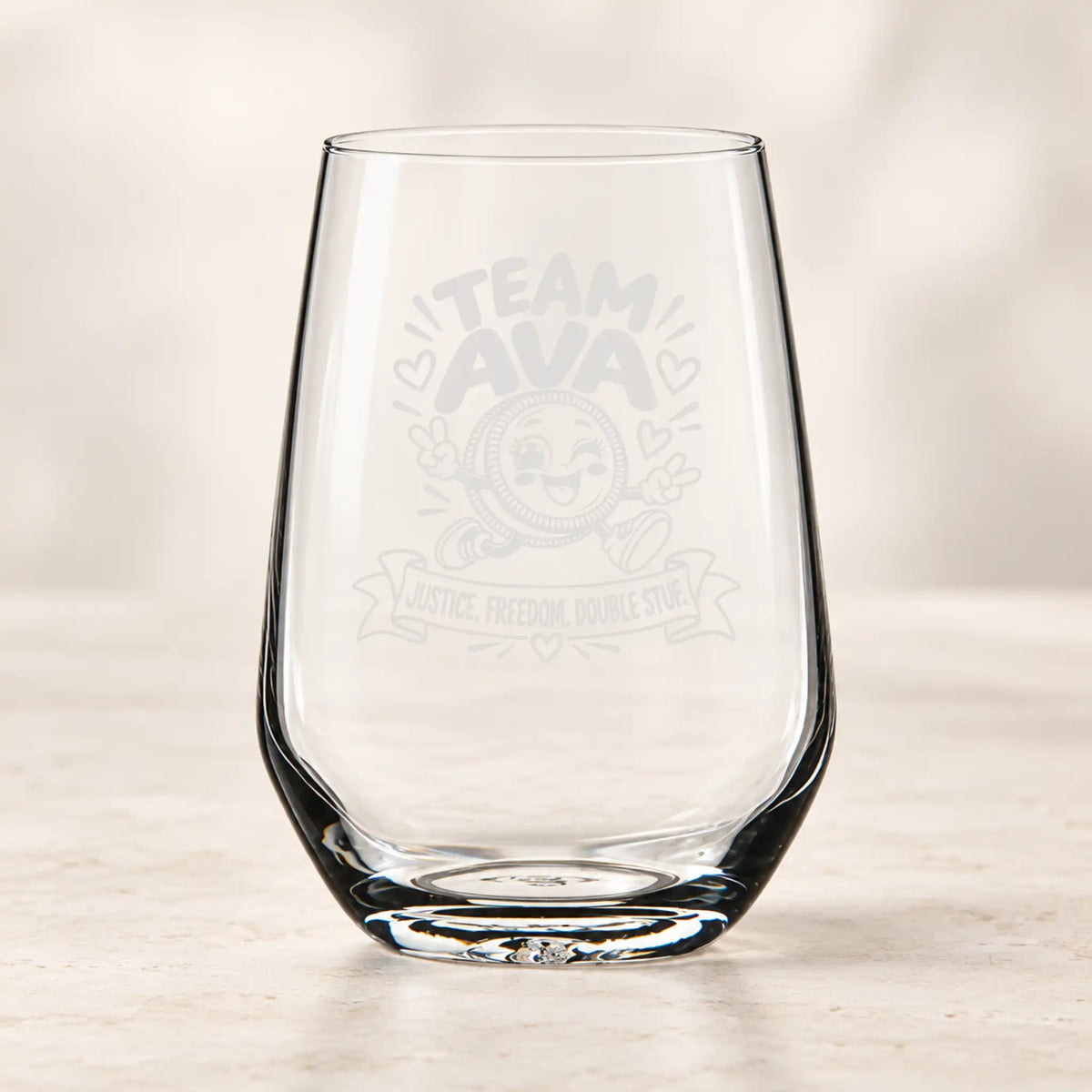 A clear stemless wine glass with a frosted gray 'TEAM AVA' graphic and emblem on the front, standing on a light marble surface against a soft beige background.
