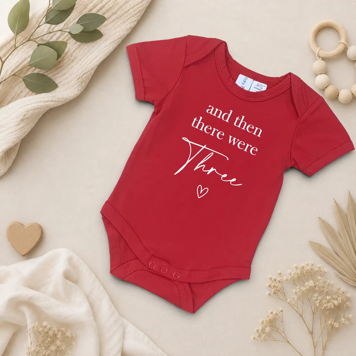 Red baby onesie with "and then there were three" on a neutral background